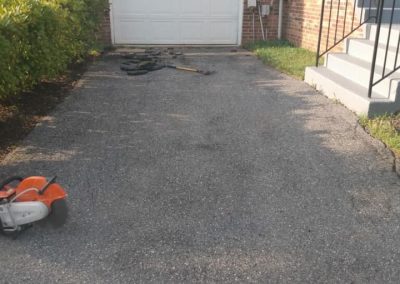 A paved driveway with tools and tires, leading to a closed garage door of a brick house, showcases the quality work of All County Paving—your go-to for expert paving solutions in Hanover MD.