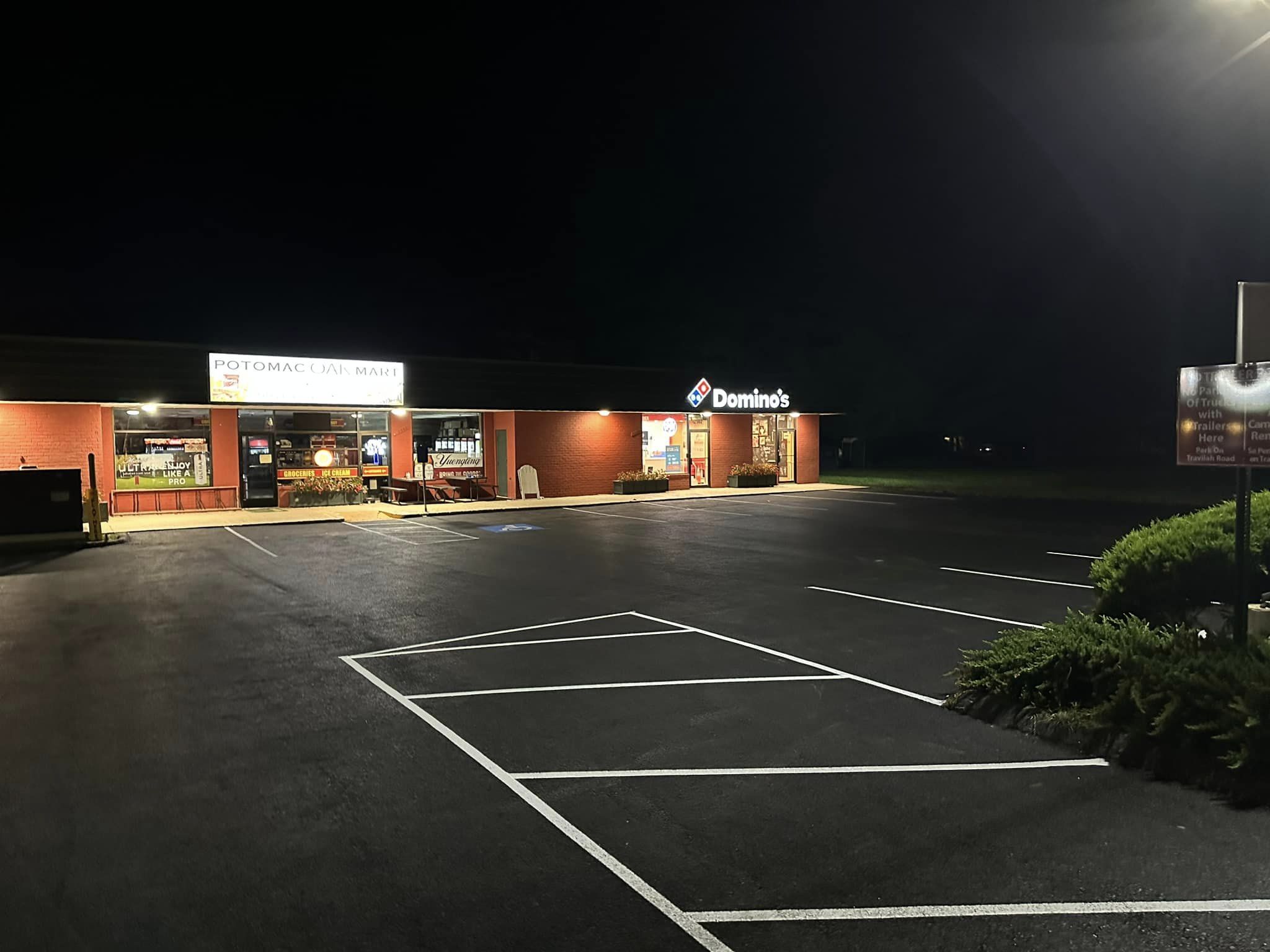 A small shopping plaza at night with Potomac Deli Market and Domino’s, their signs and parking lot lights reflecting off expert asphalt paving by All County Paving.