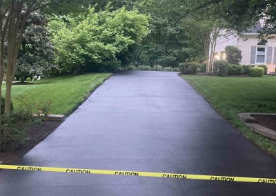 Freshly paved driveway blocked by yellow caution tape, surrounded by green trees and grass.