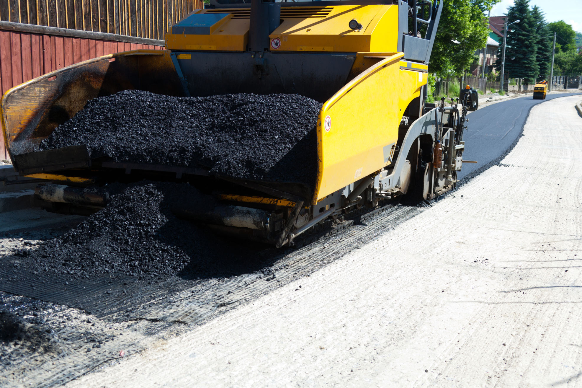 A yellow paving machine lays fresh asphalt on a road under construction on a sunny day.