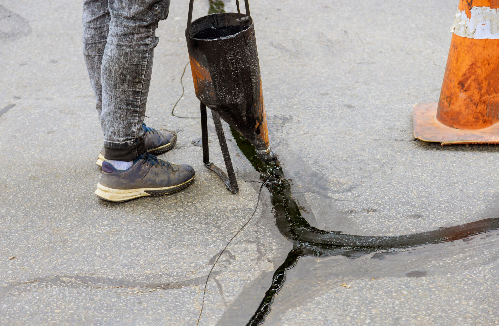 A person fills cracks in pavement with sealant near an orange traffic cone.