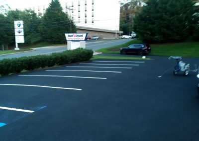 Empty parking lot with a few cars, bushes, a sign, and a large building in the background.