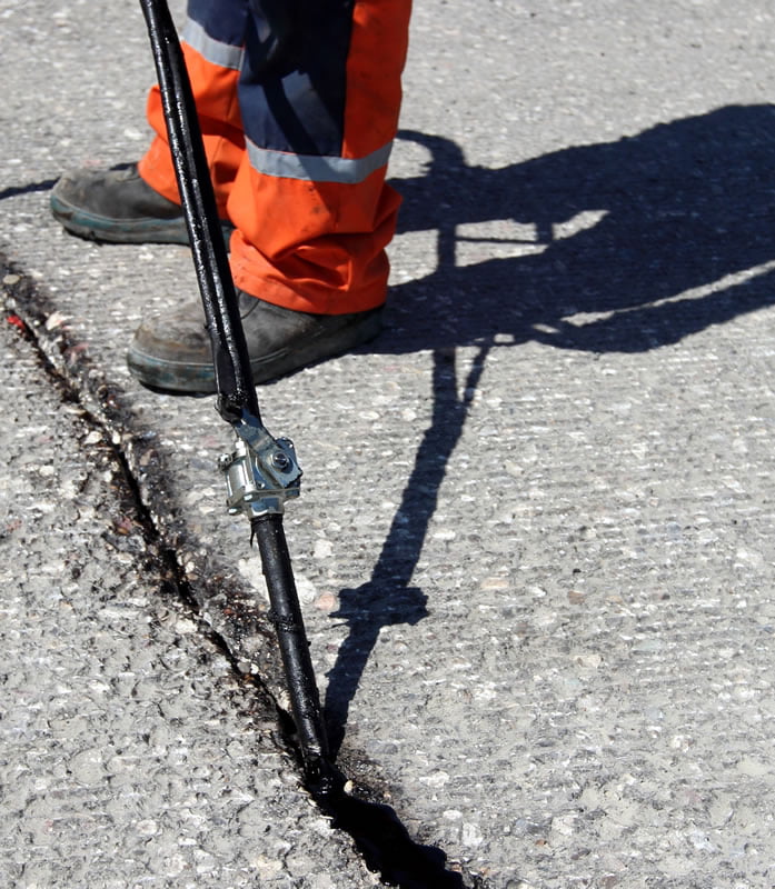 Worker in orange pants sealing a crack in asphalt with a tool on a sunny day.