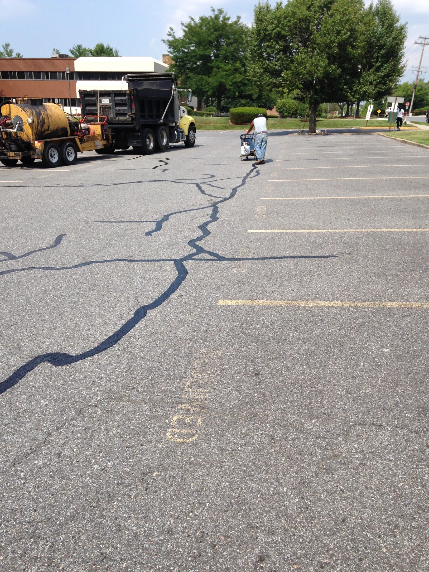 Worker sealing cracks in asphalt of an empty parking lot with equipment; truck parked nearby.