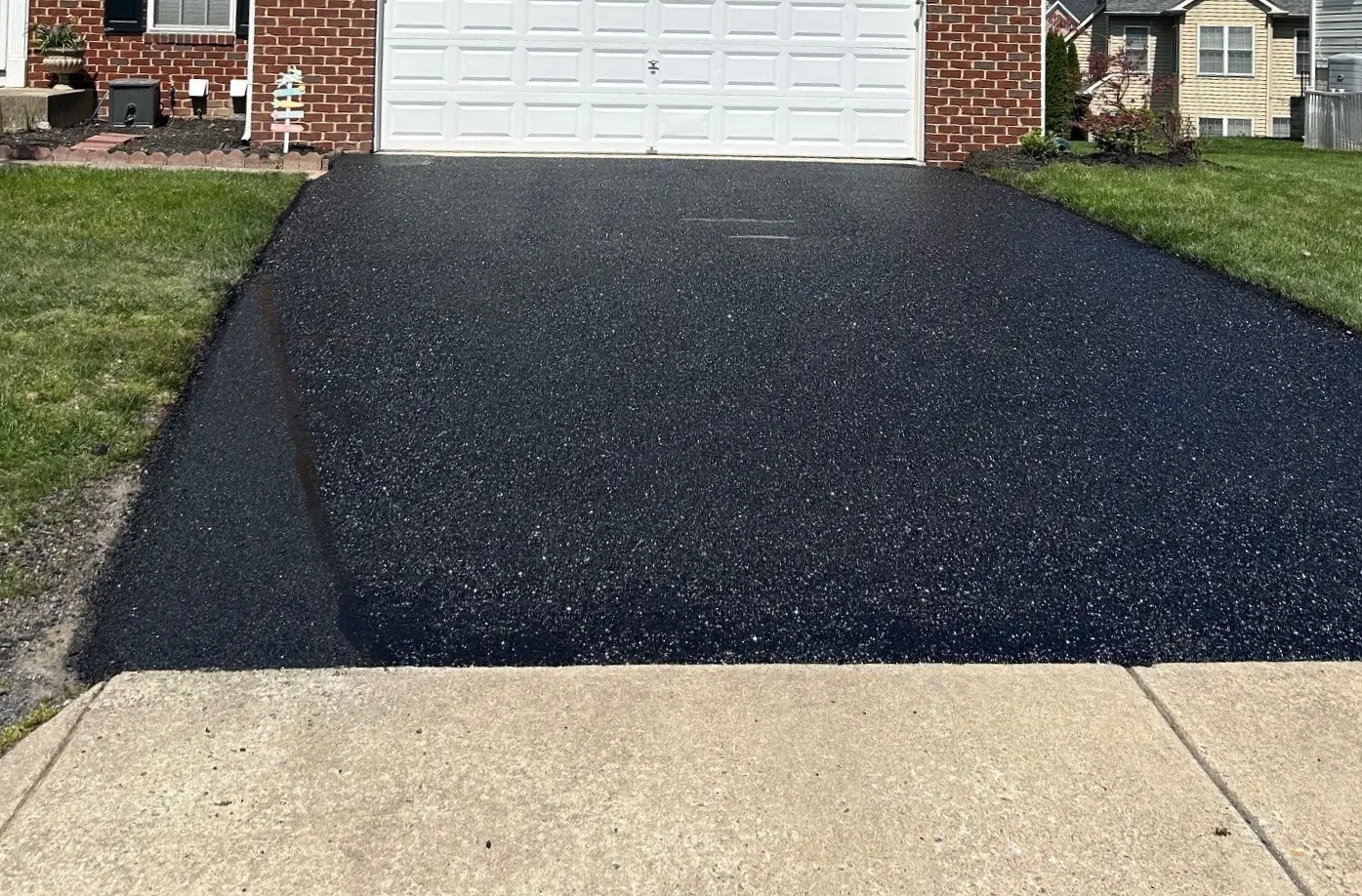 A newly sealed black asphalt driveway leads up to a white garage door of a brick house.